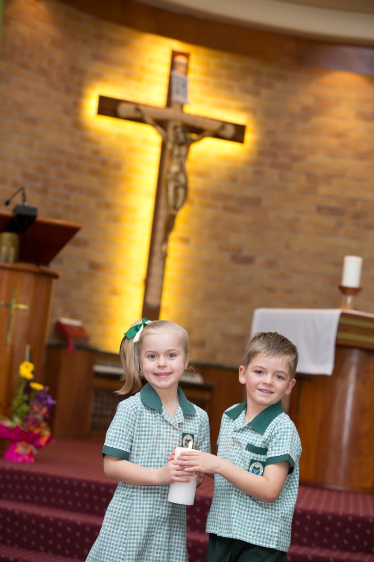 Two students in school uniforms standing at the front of a chapel holding a candle, with a large crucifix on a brick wall behind them