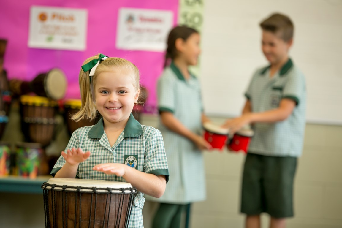 Student playing a hand drum in a music class while other students play percussion instruments in the background