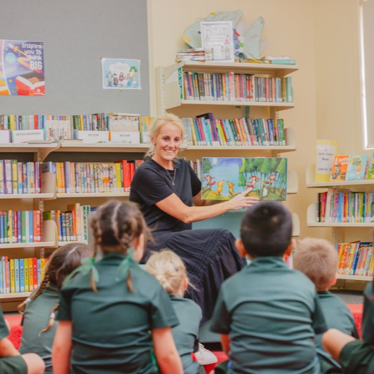 Teacher reading a picture book to seated students in a school library with bookshelves in the background