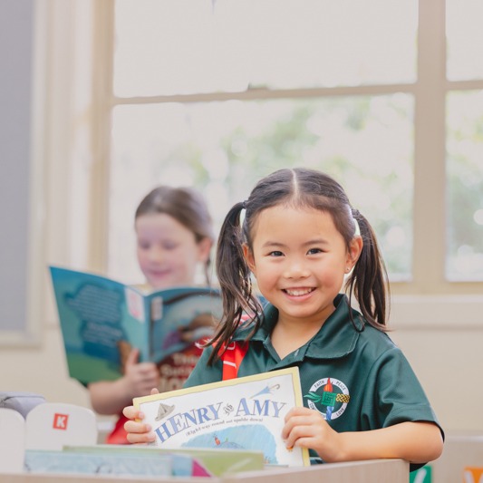 Student holding a book titled “Henry and Amy” while seated at a desk, with another student reading in the background
