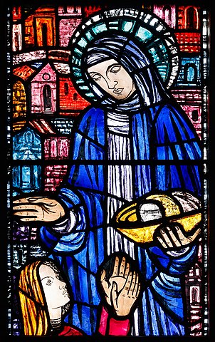 Stained glass window depicting a robed religious figure holding bread, with a halo and buildings in the background