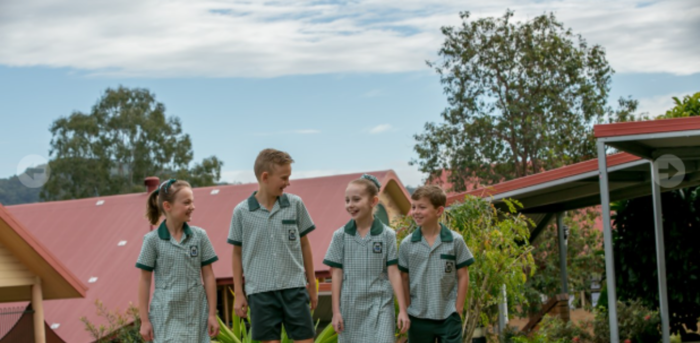 Four students in school uniforms walking together outdoors on school grounds near buildings with red roofs