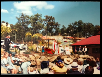 Outdoor school gathering with seated audience watching a presentation near a building with a red roof