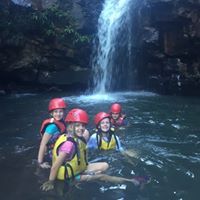 Students wearing helmets and life jackets standing in a shallow pool beneath a waterfall during an outdoor adventure activity