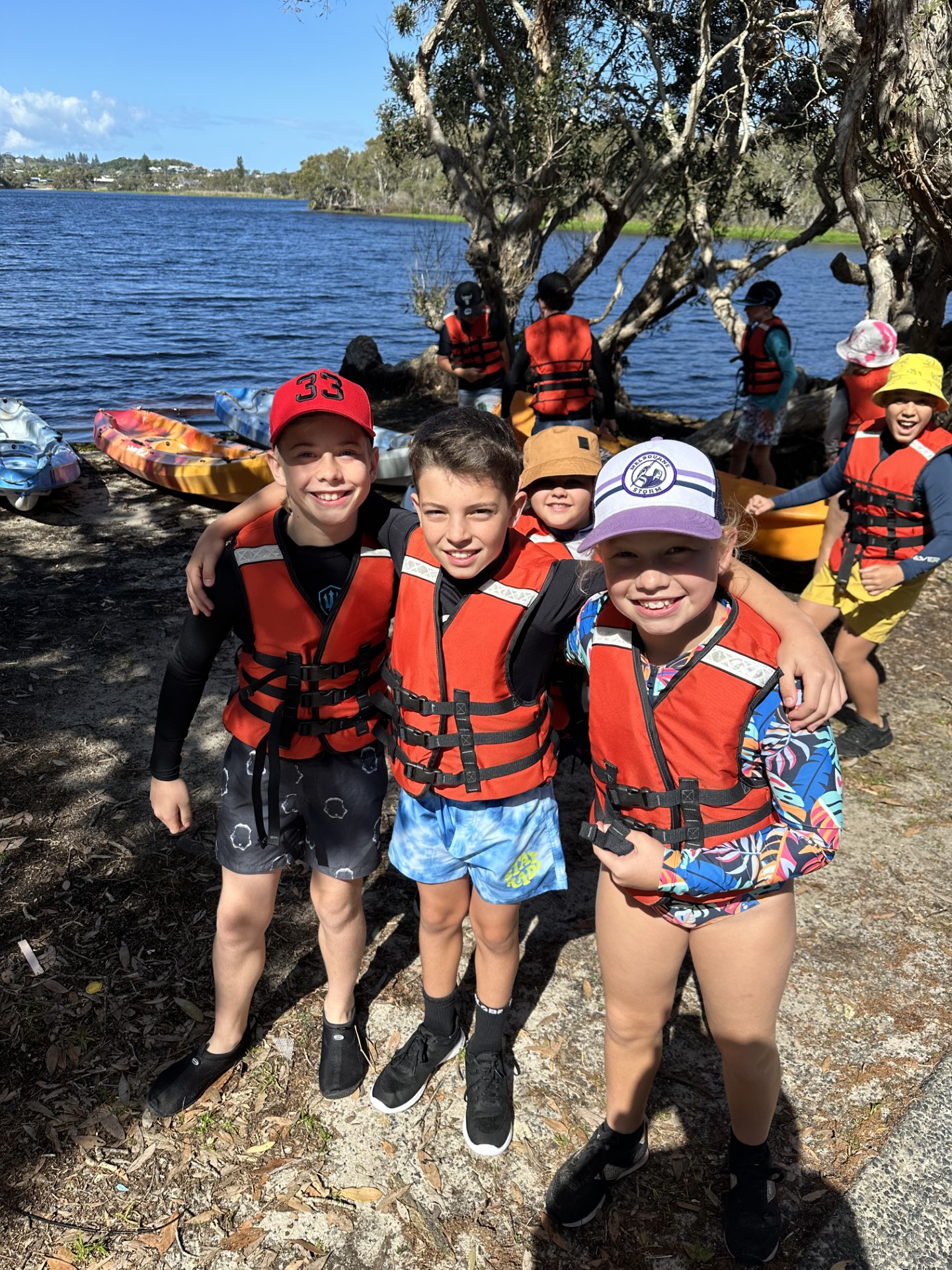 Students wearing life jackets standing together by kayaks on a lakeshore during an outdoor activity